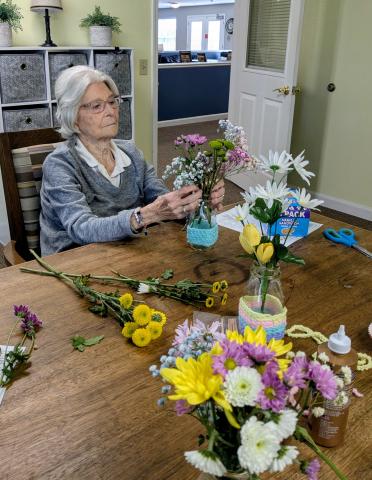 Elderly woman arranging flowers in a vase she decorated with crocheted strands.